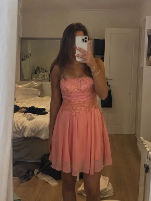 Axelbandslös kort rosa klänning - Söt axelbandslös kort klänning i rosa med vackra spetsdetaljer upptill och i midjan. Kjolen är lätt och luftig med flera lager tyg som ger ett fint fall. Perfekt för dig som vill ha en feminin och elegant look.