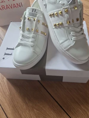 Vita Valentino Rockstud sneakers - Snygga vita sneakers från Valentino Garavani med guldiga nitar och breda band över ovandelen. Skorna har klassisk låg profil, vita snören och är tillverkade i slätt läder. Perfekta för dig som vill sticka ut med exklusiva detaljer.