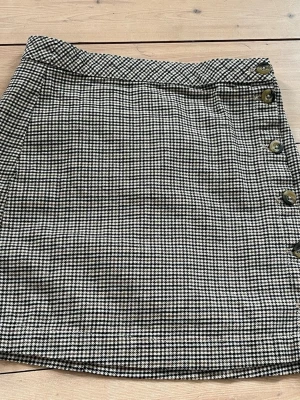 Rutig minikjol med knappar - Snygg kortkjol med svartvitt rutigt mönster och dekorativa knappar längs sidan. Kjolen har en klassisk rak passform och är perfekt att styla med en enkel topp eller stickad tröja. Använd endast en gång.