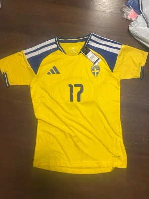 Sverige matchtröja Adidas nr 17 Gyökeres - Gul matchtröja (player-version) från Adidas för Sveriges landslag med blå detaljer, nummer 17 och namnet Gyökeres på ryggen. Tröjan har korta ärmar, blåa ränder på axlarna och landslagets emblem på bröstet. Tillverkad i lätt och ventilerande material.
