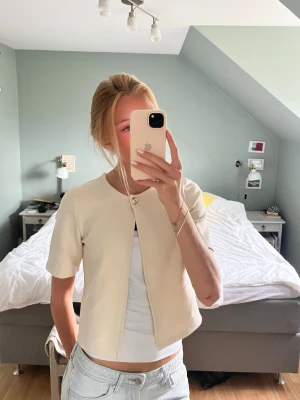 Kort cremevit kofta från Gina Tricot - Stilren, elegant kort kofta i cremevitt från Gina Tricot.💕Använd en enda gång!! Koftan har rund halsringning, korta ärmar och stängs med en dekorativ guldfärgad knapp framtill. Perfekt att bära för en ”put-together look”😇😇nypris: 300kr
