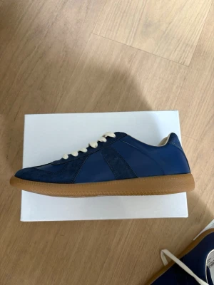 Maison Margiela blå - Snygga blå sneakers från Maison Margiela med ovandel i mocka och läder, vita skosnören och klassisk gummisula i brunt. Låg profil och stilren design som passar till många outfits.