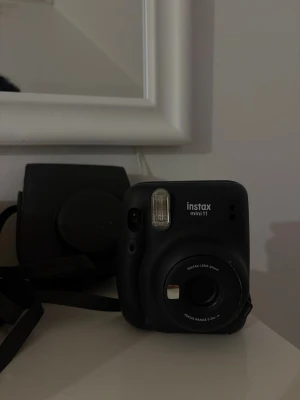 Svart Instax Mini 11 direktkamera - Säljer en svart Instax Mini 11 från Fujifilm. Kameran har en kompakt och rundad design i plast, inbyggd blixt och enkel knapp för att ta bilder direkt. Perfekt för att få ut fysiska foton direkt. Medföljer matchande svart kameraväska. perfekt nu inför sommaren för att skapa minnen. ordinarie pris 959kr
