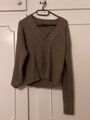 Beige ribbad kofta med v-ringning - Mysig brun kofta med v-ringning och ribbad struktur. Koftan har långa ärmar,  croppad i modellen. Perfekt att slänga över en topp för en avslappnad look.