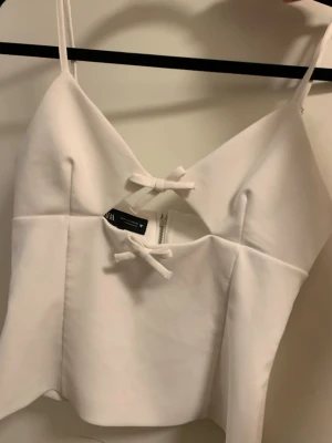Zara topp - Supersnygg vit linnetopp från Zara med smala axelband och två söta rosetter framtill. Toppen har en v-ringad skärning och lätt figurnära passform. Perfekt för sommarens alla tillfällen och enkel att styla med jeans eller kjol.
