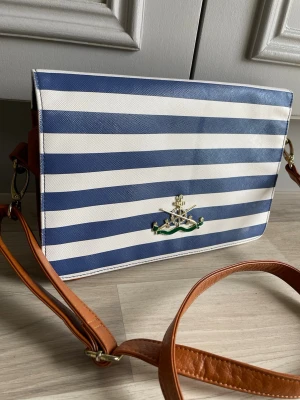 Randig axelväska från Vivienne Westwood - Snygg axelväska med blåvita ränder och brunt justerbart axelband. Väskan har ett guldfärgat Vivienne Westwood-emblem på framsidan och är gjord i ett tåligt material med läderdetaljer. Perfekt för dig som gillar en unik och trendig look.