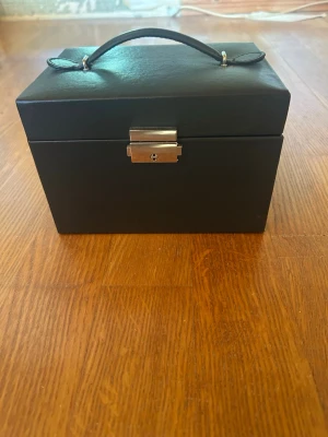 Svart smyckeskrin med spegel - Stilrent svart smyckeskrin i läderimitation med handtag och lås i silverfärgad metall. Insidan har flera fack, ringrullar och en inbyggd spegel. Vissa fack på ovansidan kan göras större. Perfekt för att organisera ringar, örhängen och halsband. Ljusbeige inredning ger en lyxig känsla.