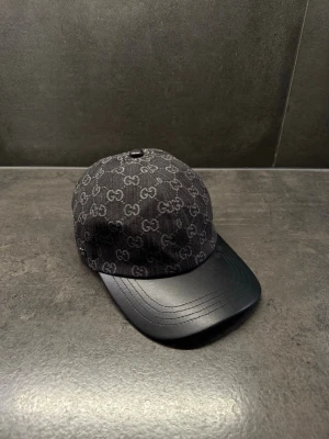 Svart GG keps med monogram - Snygg svart keps från GG med klassiskt GG-monogram över hela tyget. Skärmen är i svart läder-look som ger en exklusiv känsla. Perfekt accessoar för dig som vill ha en stilren och trendig look med lyxig touch.