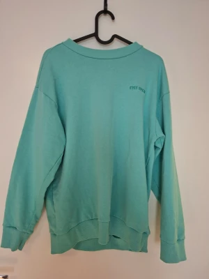 Turkos hoodie från Saligia XS - Turkos hoodie från Saligia i storlek XS. Hoodien har en avslappnad passform, ribbade muddar och nederkant samt liten text 'EYES OPEN' på bröstet. Perfekt för en chill och stilren look.