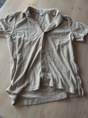 Beige frottéskjorta från East West - Beige skjorta i mjuk frotté från East West. Skjortan har korta ärmar, klassisk krage och knäppning framtill. Perfekt för en avslappnad och trendig look.