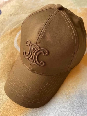 Brun keps från Celine med broderad logga - Snygg brun keps från Celine med den ikoniska broderade loggan framtill. Kepsen har böjd skärm och klassisk sexpanelsdesign. Tillverkad i ett slitstarkt tyg som ger en stilren och trendig look.