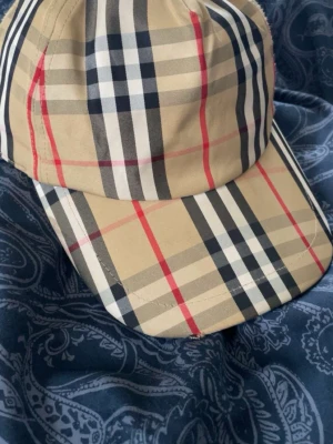 Beige keps från Burberry - Snygg keps från Burberry med klassiskt rutmönster i beige, svart, vitt och rött. Märket 'Burberry London England' är broderat på baksidan. Finns en liten defekt vid framsidan som knappt syns 