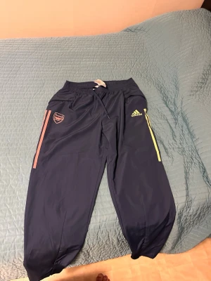 Arsenal träningsset från Adidas - Snyggt Arsenal träningsset från Adidas med marinblå byxor med rosa och gula detaljer samt klubbmärke. Matchande ljusgul jacka med blå och rosa ränder, huva och dragkedja. Perfekt för fotbollsträning eller sportiga dagar. s på tröjan men sitter som M byxorna är L