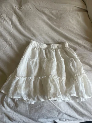 Vit volangkjol från B Sister XS - Supersöt vit kortkjol från B Sister i storlek XS. Kjolen har flera volanger och ett diskret broderat mönster, samt resår i midjan för extra komfort. Perfekt för en fräsch och somrig look!