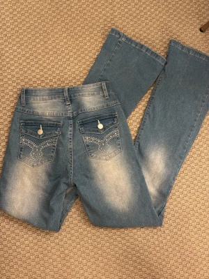 Blå bootcut jeans med dekorerade fickor - Snygga ljusblå bootcut jeans med slitningar och dekorerade bakfickor med vita sömmar och knappar. Jeansen har klassisk femficksdesign och är tillverkade i denim. Perfekta för en avslappnad och trendig look.