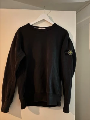 Svart sweatshirt från Stone Island - Cool svart sweatshirt från Stone Island med klassisk rund hals och ikonisk logotyp-patch på vänster ärm. Tröjan har en avslappnad passform och är tillverkad i mjukt bomullsmaterial. Perfekt för en stilren och trendig look.