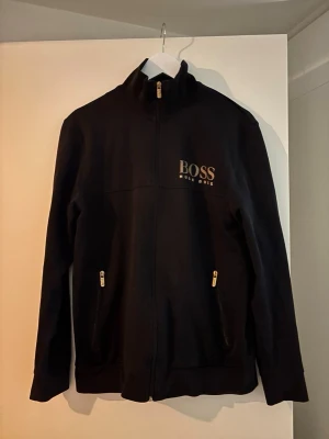 Svart zip-hoodie från Hugo Boss - Svart hoodie med dragkedja från Hugo Boss. Stort BOSS-tryck i vitt på bröstet och två fickor med guldfärgade dragkedjor. Hög krage och stilren design, perfekt för en clean och snygg look.