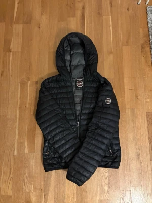 Svart pufferjacka från Colmar - Snygg svart pufferjacka från Colmar med huva och dragkedja framtill. Jackan har två fickor med dragkedja och Colmar-logga på ärmen. Lätt vadderad och quiltad design som håller värmen under kalla dagar. Tyvärr en trasig dragkedja men jackan går att stänga fullt normalt.