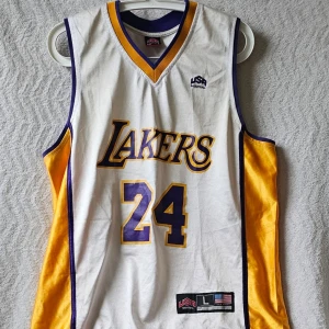 Lakers basketlinne nr 24 - Snyggt basketlinne från USA Basketball med Lakers-tryck och nummer 24. Linnet är vitt med lila och gula detaljer, v-ringad hals och broderade loggor. Perfekt för basketträning eller som streetwear. Materialet är lätt och andas bra.