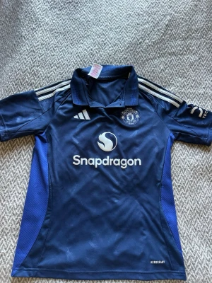 Manchester United tränings t-shirt blå - Snygg blå Manchester United tränings t-shirt från Adidas med Snapdragons logga på bröstet. Tröjan har vita ränder på axlarna, klubbmärke på vänster bröst och är tillverkad i lätt och ventilerande material (AEROREADY). Perfekt för fotbollsträning.