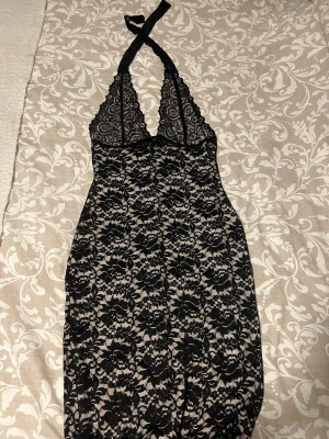 Svart spets klänning med halterneck - Snygg klänning i svart spets med halterneck och djup urringning. Underklänning i beige för en kontrast mot spetsen. Tillverkat i 95% polyester och 5% elastan för en mjuk och stretchig känsla. Perfekt för en lyxig och bekväm käll.