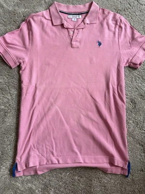 Rosa pikétröja från U.S. Polo Assn - Säljer en klassisk rosa pikétröja från U.S. Polo Assn med blå broderad logga på bröstet. Tröjan har krage, knappar framtill och korta ärmar. Perfekt till jeans eller shorts för en avslappnad look.