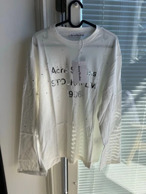 Vit långärmad topp Acne Studios - Vit långärmad topp från Acne Studios med silverfärgad text och små glittriga detaljer framtill. Rund halsringning och rak passform. Perfekt för dig som gillar stilrena plagg med en twist.