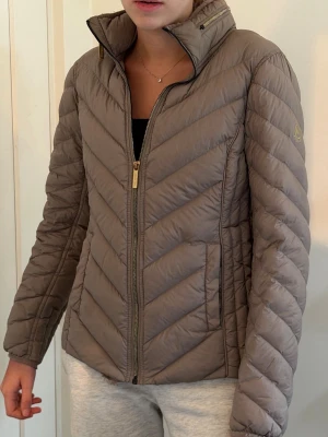 Taupe pufferjacka från Michael Kors - Snygg taupefärgad pufferjacka från Michael Kors med guldfärgade dragkedjor och hög krage. Jackan har ett quiltat mönster i diagonala linjer och diskret logga på ärmen. Perfekt för kalla dagar och riktigt skön att bära. Nypris 2269kr 