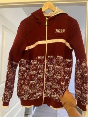 Vinröd hoodie från Hugo Boss - Snygg vinröd hoodie från Hugo Boss med vit dragkedja och huva. Hoodien har ett vitt band över bröstet och ett coolt mönster med BOSS-loggor över hela nedre delen och ärmarna. Perfekt för dig som vill sticka ut med stilren streetwear.