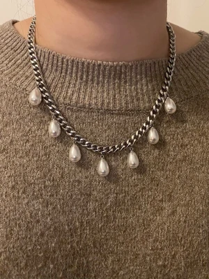Kedjehalsband med droppformade pärlor - Snyggt halsband med en grov silverfärgad kedja och sju droppformade vita pärlor som hänger längs kedjan. Halsbandet har en modern och edgy känsla, perfekt för att lyfta din outfit med en cool detalj. Materialet är rostfritt och pärlorna.