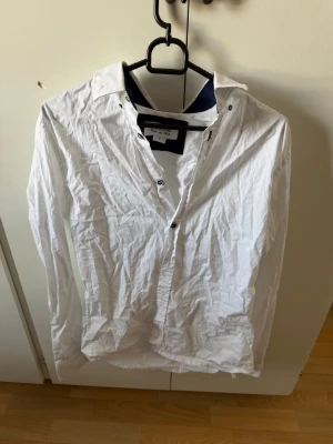 Vit skjorta från Missone med knappar - Klassisk vit skjorta från Missone med lång ärm och knappar framtill. Skjortan har en stilren design med mörkblå detaljer på insidan av kragen. Perfekt för dig som gillar en enkel men snygg look.