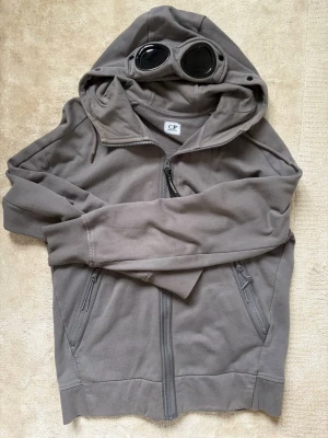 Grå hoodie med goggles från C.P. Company - Säljer en grå hoodie från C.P. Company med dragkedja och två fickor framtill. Huvan har inbyggda goggles som ger en unik och cool look. Perfekt för dig som vill sticka ut med streetwear-stil.