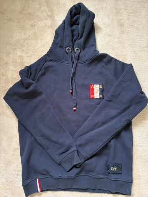 Mörkblå hoodie från Ace - Mörkblå hoodie från Ace med broderad logga på bröstet i rött, vitt och grått. Huva med dragsko och metallöljetter, samt ribbade muddar vid ärmslut och nederkant. Liten detalj i rött och vitt vid midjan. Perfekt för en avslappnad stil.