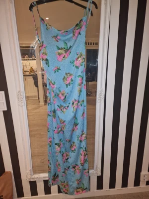 Adoore blå långklänning med smala band size 40 - Långklänning i ljusblått med blommigt mönster i rosa, grönt och gult. Klänningen har smala axelband och en rak, ledig passform. Perfekt för sommarens varma dagar och har en mjuk, lätt känsla i materialet.