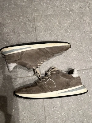 Grå mockarunners från Philippe Model - Snygga sneakers/runners i grå mocka från Philippe Model med vita detaljer och klassisk låg siluett. Skorna har vita snören, vit sula med grå och svarta inslag samt diskret logga på sidan. Perfekta för en stilren och avslappnad look.