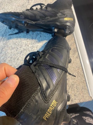 Adidas Predator svarta fotbollsskor - Säljer ett par svarta Adidas Predator fotbollsskor med klassisk design och guldtext på hälen. Skorna har snörning, syntetmaterial och stickad strumpliknande krage för extra stöd. Perfekta för dig som vill dominera på planen. Ser ut helt nya! Använda o gott skicka, skorna har fortfarande mycket att ge! 