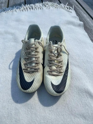 Nike Air Zoom Mercurial fotbollsskor - Säljer ett par vita Nike Air Zoom Mercurial fotbollsskor med stora svarta Nike loggor på ovansidan. Skorna har snörning och är tillverkade i syntetmaterial för lätt vikt och bra grepp på planen. Perfekta för dig som vill ha snabbhet och kontroll på fotbollsplanen.