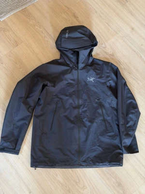 Svart SL regnjacka från Arc'teryx - Snygg svart regnjacka från Arc'teryx med huva och vattentäta dragkedjor. Jackan har två stora fickor framtill och är tillverkad i ett slitstarkt, vattenavvisande material. Perfekt för regniga dagar och outdoor-äventyr. Size runs small so XL = L