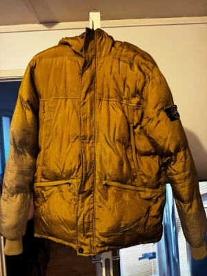 Stone Island gul pufferjacka med huva - Grym gul pufferjacka från Stone Island med broderad logga på ärmen och huva. Jackan har två snedställda fickor framtill och dragkedja med knappslå. Perfekt för kalla dagar och riktigt snygg streetstil. Materialet är glansigt och ger jackan en exklusiv känsla.