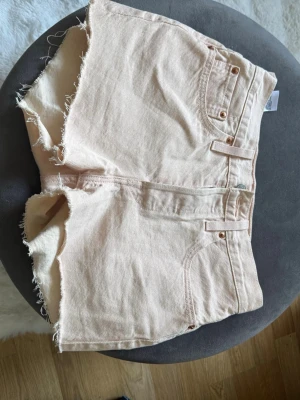 Beige jeansshorts från Levi's - Säljer ett par snygga rosa jeansshorts från Levi's med råa kanter och klassisk femficksdesign. Shortsen har bälteshällor, perfekt för en chill sommarlook. Levi's-logga på bakfickan och slitna detaljer för extra edge.