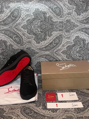 Svarta Christian Louboutin sneakers - Snygga svarta sneakers från Christian Louboutin med klassisk röd sula. Skorna har en låg profil, svarta skosnören och ovandel i mocka. Perfekta för dig som vill sticka ut med exklusiv design och ikoniska detaljer.