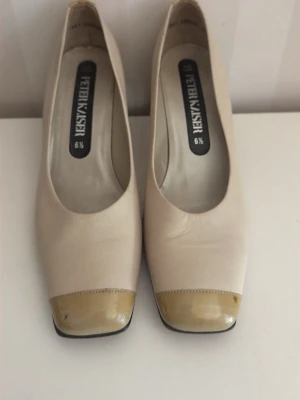 Beige pumps med lackad tå Peter Kaiser - Stilrena pumps från Peter Kaiser i beige läder med fyrkantig tå och glansig, ljusbeige lackad tådetalj, liten fläck på ena tån.Klassisk modell med låg klack och diskret branding på innersulan. Perfekta för dig som gillar en tidlös och elegant look.