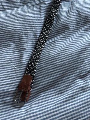 Svartvitt flätat bälte med läderdetaljer - Snyggt flätat bälte i svart och vitt med bruna läderdetaljer vid spännet. Bältet har ett klassiskt metallspänne och passar perfekt till jeans eller chinos. Den flätade designen gör det enkelt att justera längden efter behov.