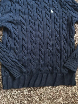 Mörkblå kabelstickad tröja Polo Ralph Lauren - Snygg mörkblå kabelstickad tröja från Polo Ralph Lauren med klassisk vit logga broderad på bröstet. Tröjan har rund halsringning, långa ärmar och ribbade muddar vid ärmslut och nederkant. Perfekt för en stilren och tidlös look.