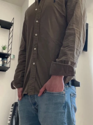 Olivgrön skjorta från Ralph Lauren - Olivgrön skjorta från Ralph Lauren med klassisk krage och knäppning framtill. Skjortan har långa ärmar och en liten broderad logga på bröstet. Tillverkad i bomull och passar perfekt till jeans eller chinos.