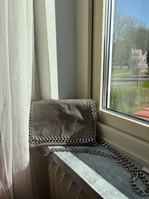 Grå  axelväska  med kedja - Snygg grå axelväska  i veganskt läder med grov struktur. Väskan har silverfärgad kedja som axelrem och dekor runt kanterna samt magnetlås och innerfack med dragkedja. Perfekt för dig som gillar trendiga accessoarer. Helt felfri och rymlig!