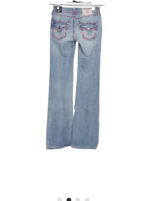 Ljusblå bootcut jeans True Religion - Ljusblå bootcut jeans från True Religion med rosa kontrastsömmar och dekorativa bakfickor med lock och knappar. Klassisk femficksmodell i denim med låg midja. Perfekta för en avslappnad och trendig look.