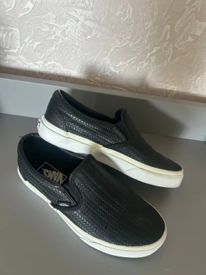 Svarta slip-on Vans sneakers - Säljer ett par svarta slip-on Vans sneakers med vit sula. Ovandelen har ett diskret vävt mönster i material som liknar konstläder. Klassisk Vans-logga på sidan och bekväm låg profil. Perfekta för en avslappnad streetwear-look.