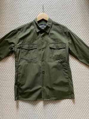 Olivgrön overshirt från Mads Nørgaard - Stilren olivgrön overshirt från Mads Nørgaard med klassisk krage, två bröstfickor med lock och knappar framtill. Tillverkad i ett lätt bomullstyg som passar perfekt för lager på lager. Enkel och clean design som funkar till det mesta.