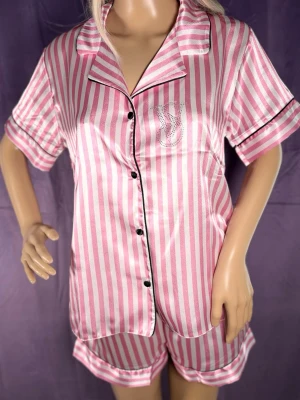Rosa randig satinpyjamas med kort ärm - Snygg pyjamas i glansigt satinmaterial med rosa och vita ränder. Kortärmad skjorta med svarta knappar och svart kant, samt matchande shorts. Bröstet har ett glittrigt VS-tryck. Perfekt för mysiga kvällar hemma.